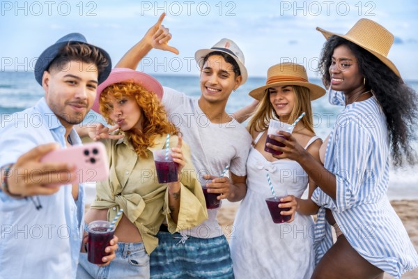 Group of cheerful young multi ethnic friends in summer clothes taking selfie with smartphone and drinking cocktails on the beach