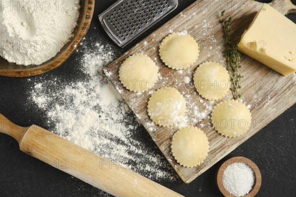 Fresh pasta dough is rolled out on a wooden board surrounded by flour, cheese, and herbs. Round pasta shapes are carefully placed, ready for cooking or stuffing.