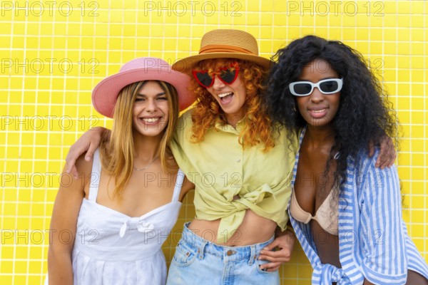 Three cheerful diverse women with colorful hats and sunglasses are posing together against a bright yellow tiled wall, representing summer joy and friendship