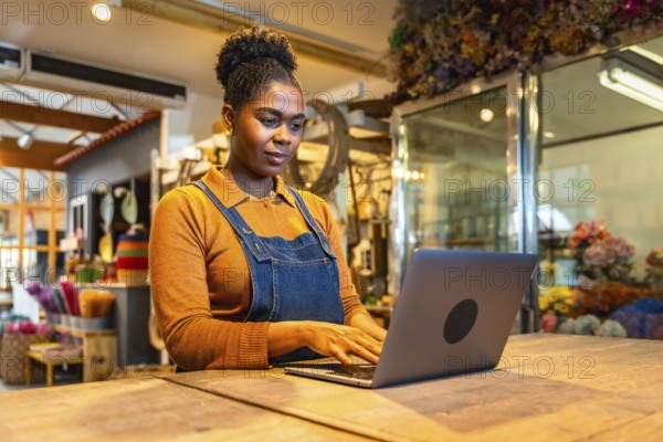 Young florist managing online orders using laptop in her flower shop, surrounded by colorful flowers and plants