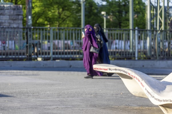 New seating on Norbert-Burger-Platz at the end of the Hohenzollern Bridge on the right bank of the Rhine. Inscription in various languages: What do you want in Cologne? Two Muslim women with burqas. Cologne, North Rhine-Westphalia, Germany