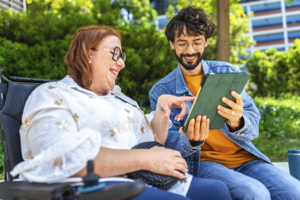 Man and a woman are using a digital tablet in a park, she is sitting in a wheelchair and they are both smiling