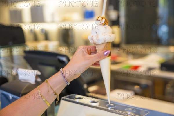 Womans hand adorned with bracelets, holding a delicious ice cream cone topped with crunchy nuts, enjoying a treat at a gelateria