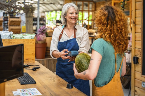 Smiling greengrocer holding pos terminal while customer paying with contactless payment for groceries in a small local shop