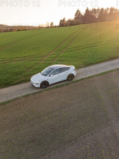 A white car on a country lane, framed by green fields and a warm evening sun on the horizon, Tesla, New Model Y, Black Forest, Germany