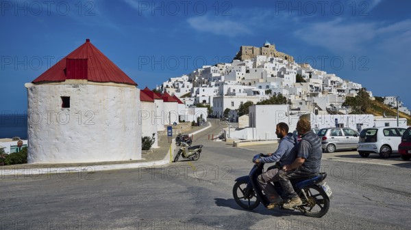 Two people on motorbike in front of white village and windmills on hill, Querini Castle, Chora, Astypalea, Dodecanese, Greek Islands, Greece