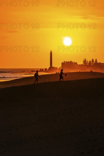 Tourists walking on the maspalomas dunes at sunset, with the maspalomas lighthouse in the background, gran canaria, canary islands, spain