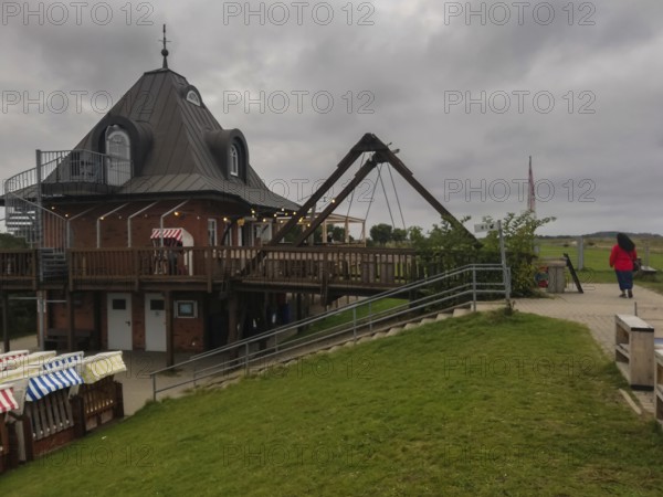 Wooden building with outdoor terrace under a cloudy sky, a person walks past Strandbude Ostseeinsel Fehmarn Fehmarnsund Germany Putgarden