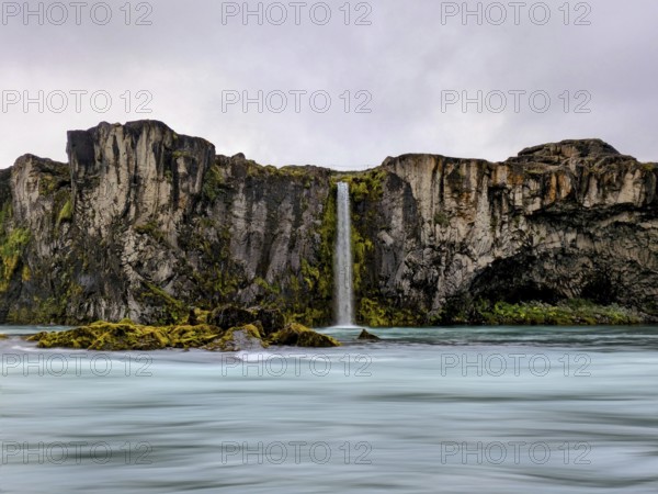 Vibrant waterfall cascading down moss-covered cliffs under a cloudy sky, Icelandic scenery. Iceland