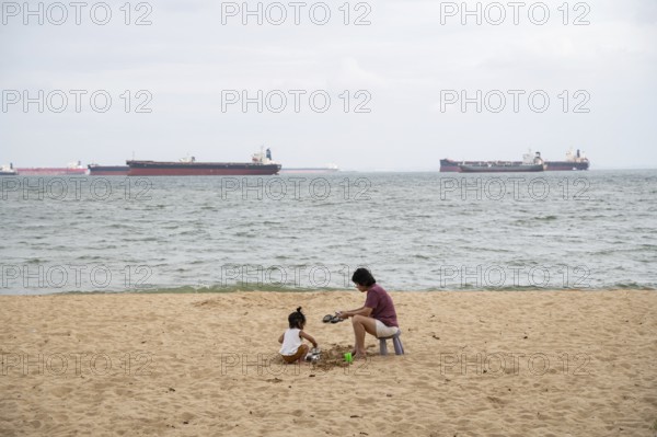 31.07.2023, Singapore, Republic of Singapore, Asia, A woman and a small child sit in the sand on the beach of East Coast Park while container ships can be seen on the water in the background