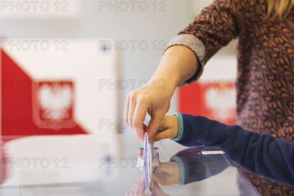 Woman's and kid's hands puting a card with a vote to the ballot box during parliamentary elections. In the backround polish arms and flag