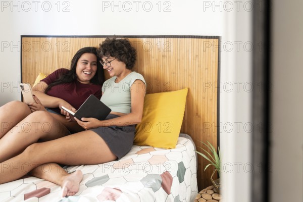 A joyful couple relaxes on a cozy bed. They are sharing a moment, with one reading a notepad and the other using a smartphone, showcasing a casual, happy indoor scene