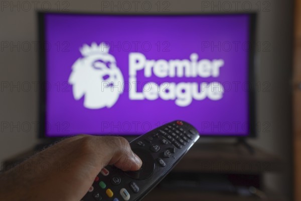 May 14, 2024, Brazil. In this photo illustration, a hand holding a TV remote control in front of the Premier League logo on a TV screen