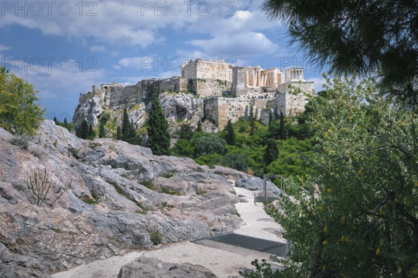 View of Acropolis hill from Areopagus hill in summer daylight with great clouds in blue sky, Athens, Greece. UNESCO world heritage. Propylaea entrance, Agrippa's monument, Parthenon. Fresh green foliage and stone and rocks of hilltop