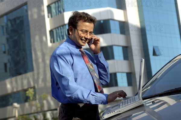 Young Indian Executive with tie working on laptop and cell phone on car bonnet in front of modern building