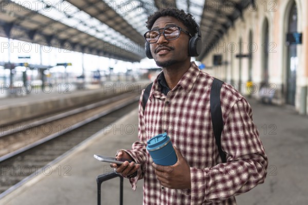 Traveler with headphones and backpack holding smartphone and reusable coffee cup is waiting for train at railroad station