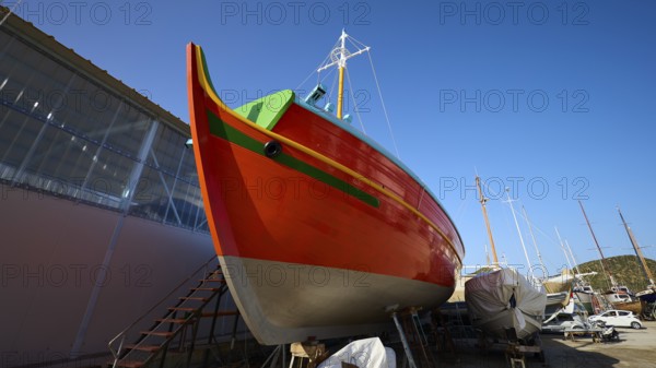 Side view of an orange-coloured boat in a shipyard under a blue sky, Diakofti boatyard, new construction of a coloured wooden boat, Diakofti, Patmos, Dodecanese, Greek Islands, Greece