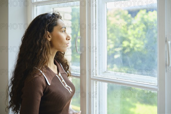 A young woman with long, curly hair is looking out a large window. Bright sunlight fills the room, and the lush greenery outside creates a serene atmosphere. She appears contemplative and calm