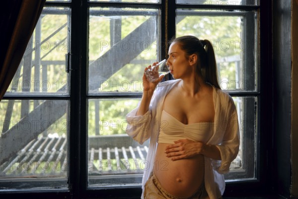 A pregnant woman stands by a large window in her home, sipping water from a glass. She appears relaxed and peaceful, enjoying a tranquil moment as natural light filters through the glass