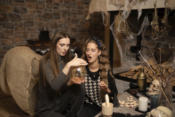 Two women sit on a cozy floor, focused on a swirling crystal ball amidst a warm, inviting atmosphere. The setting features a rustic fireplace, candles, and decorative elements for a mystical evening