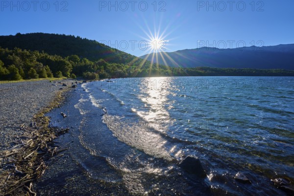 A tranquil lake with sunbeams hitting the water and shore, surrounded by forested mountains under clear skies, summer, Lake Rotoiti, Saint Arnaud, Tasman Region, South Island, New Zealand