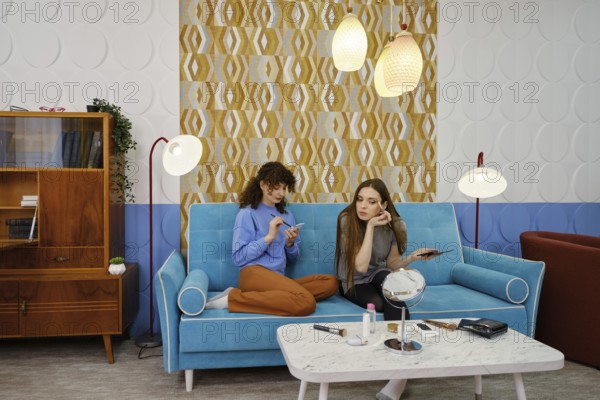 Two young women are engaged in a casual conversation about makeup while sitting on a stylish blue couch in a living room