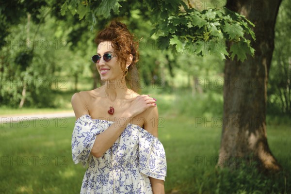 A woman with curly hair stands in a vibrant park, wearing a stylish off-shoulder dress and sunglasses. She smiles warmly while enjoying the sunny atmosphere amidst the greenery