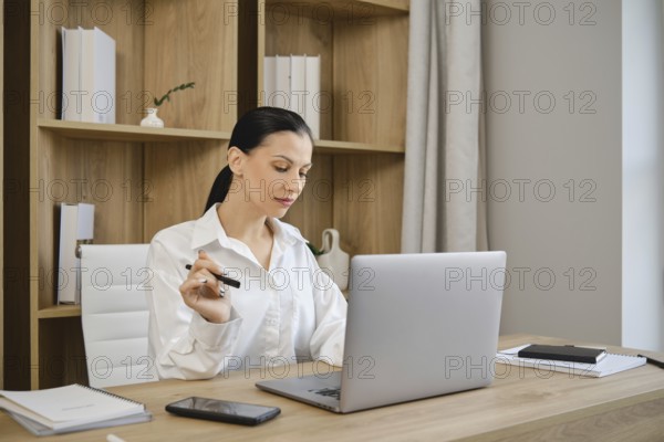 A young woman sits at a clean wooden desk focused on her laptop. She holds a pen in one hand, surrounded by notebooks and a smartphone. The minimalist office features light-colored walls and shelves