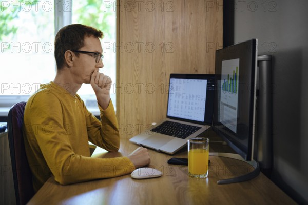 A middle-aged man sits at a wooden desk in a home office, focused on a computer screen displaying data visualisations