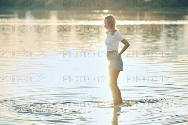 A young woman in a white top and denim shorts stands in shallow water during sunset. The golden sunlight reflects on the water's surface, creating a serene atmosphere as she enjoys the moment