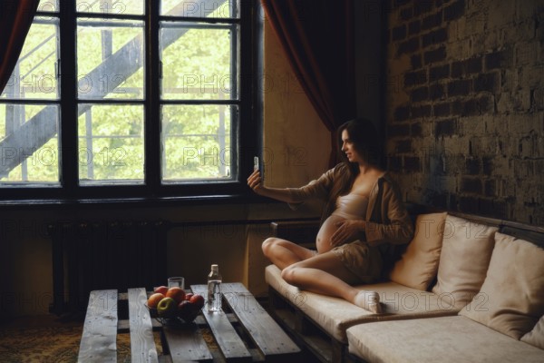 A pregnant woman sits on a comfortable couch at home, casually taking a selfie with her phone. Sunlight streams through the window, illuminating her serene expression
