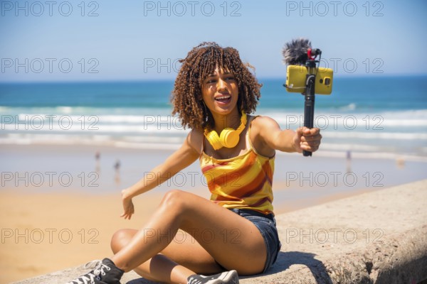 Young woman vlogger recording video blog on sunny beach using mobile phone, gimbal and microphone