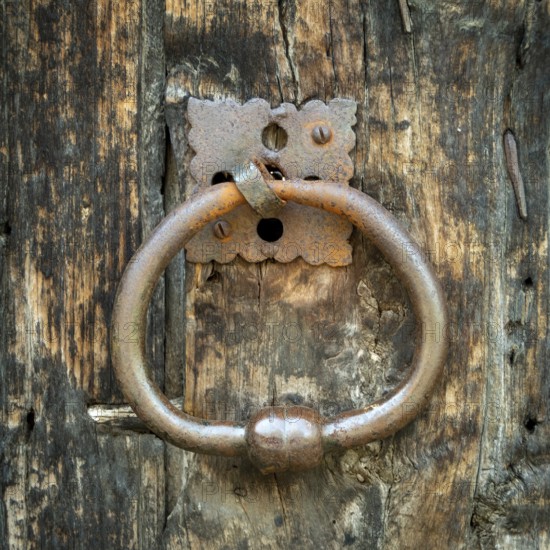 A round metal door knocker hangs on an aged wooden door, showcasing its rustic texture and worn paint. The contrasting materials highlight the history and character of the entrance