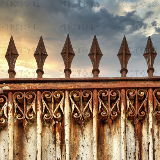 A rusty ornate gate stands tall, featuring intricate designs and pointed spikes. The backdrop shows a dramatic sky adorned with clouds during sunset, casting a warm glow on the metalwork