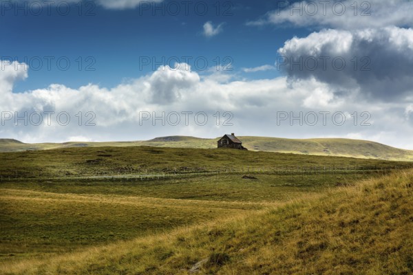 Ruined stone cottage stands alone on a grassy hillside under a partly cloudy sky in a remote countryside setting. Cezallier. Puy de Dome. Auvergne Rhone Alpes. France