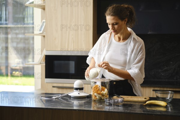 A woman is pouring milk into blender for banana and sea buckthorn smoothies in a modern kitchen