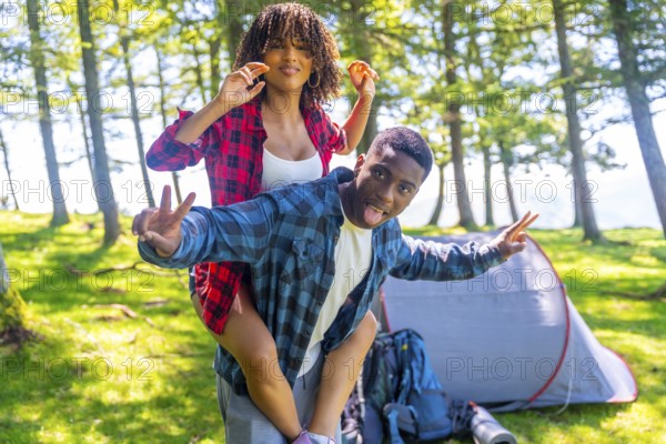 Young couple having fun piggybacking and making funny faces near their tent in a forest campground