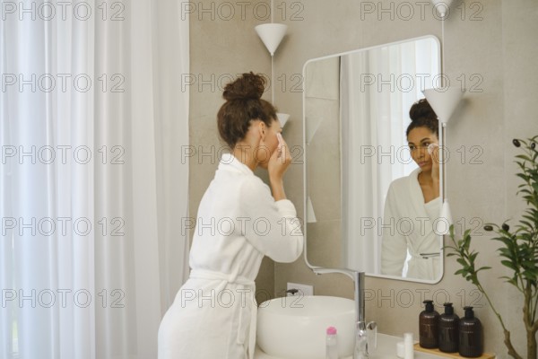 A woman in a white bathrobe is applying skincare products in a contemporary bathroom. She stands near a large mirror, focusing on her reflection while a plant adds a touch of nature