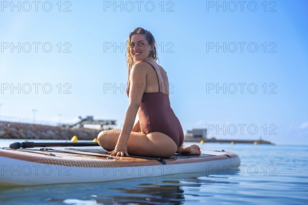 Smiling woman sitting on a paddleboard in calm blue waters, enjoying a sunny summer day at the beach. Clear sky and distant shoreline in the background