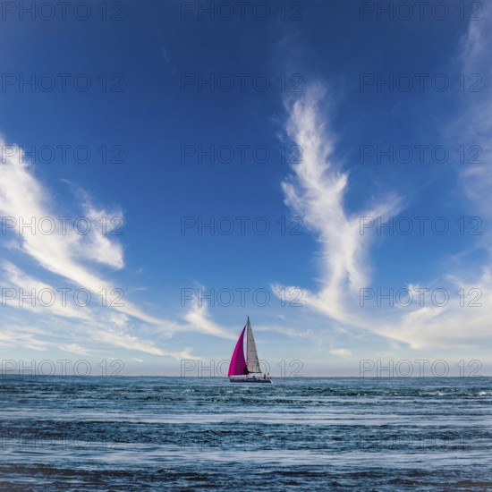 A sailboat glides across the Atlantic Ocean near Brittany, France. Its colorful sails contrast against the deep blue of the water and the striking clouds overhead