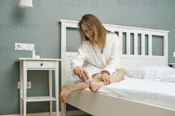 A woman in a bathrobe sits on the edge of a bed using exfoliator on her feet. The room features a calming green wall and simple furniture, creating a relaxing atmosphere for self-care