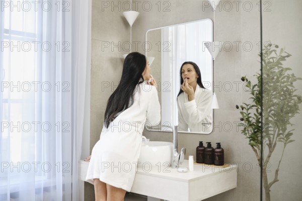 A woman in cotton robe is washing off lipstick in a bathroom, standing in front of a large mirror