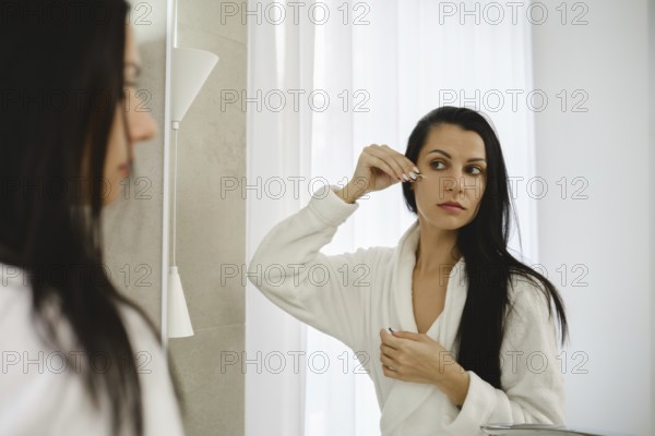 A woman in a white robe looks into a mirror while applying a beauty treatment to her under eye skin. Natural light filters through the window, creating a serene environment for her morning routine