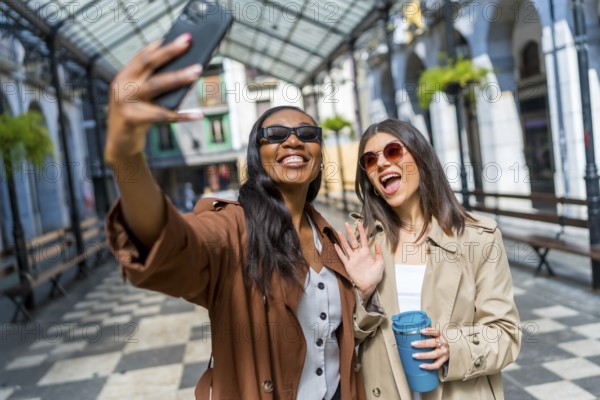 Two cheerful businesswomen taking a selfie with a smartphone and waving at the camera while walking in the city