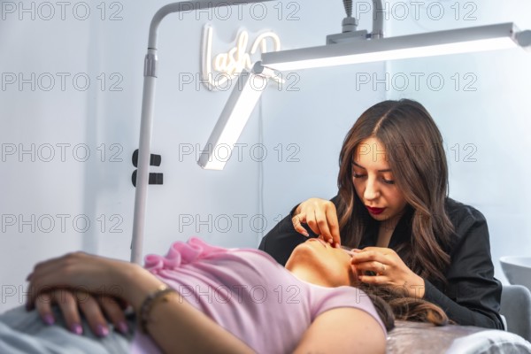 Beautician applying false eyelashes to a client lying on a bed in a beauty salon, illuminated by a professional lamp