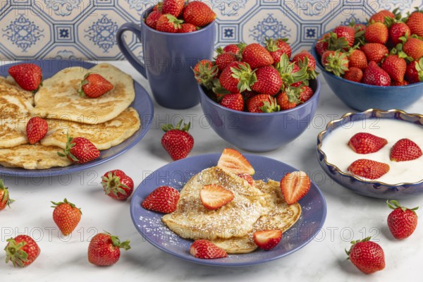 Pancakes sprinkled with icing sugar and fresh strawberries on plates next to yoghurt, blue crockery