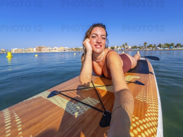 Young woman lying on a paddle surf board, taking a selfie and enjoying a sunny summer day at the beach