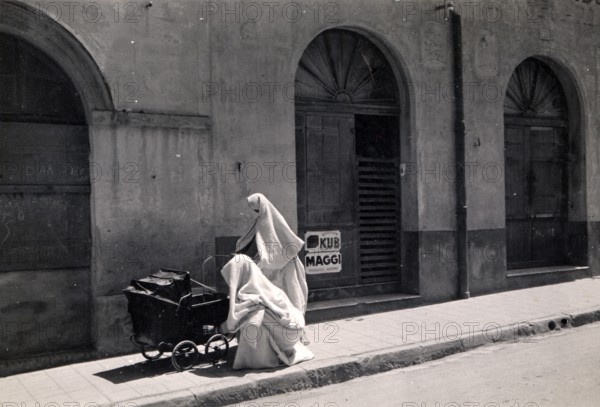 Two Arab women with prams, 1910s, A veiled person sits next to a pram in front of an old building façade, Historical photo
