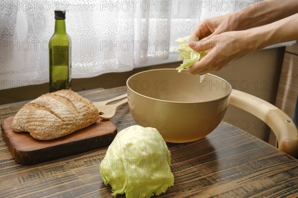 Hands are tearing fresh lettuce over a bowl while a loaf of bread and a bottle of olive oil sit nearby. Sunlight filters through a curtain, creating a warm atmosphere in the kitchen