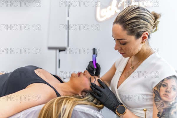 Beautician performing microblading treatment, applying permanent makeup on eyebrows of client lying on couch in beauty salon